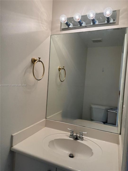 Vanity featuring an oval undermount sink, integrated solid-surface countertop, gold-toned wall-mounted towel ring, four-bulb vanity light fixture, and a wall-to-wall mirror