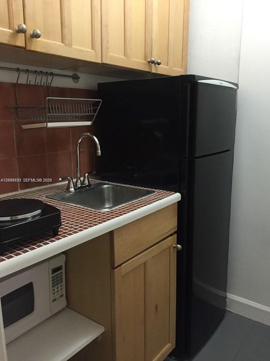 Kitchenette featuring wood-finish cabinetry, tiled countertop, stainless steel sink with metal faucet, and black refrigerator