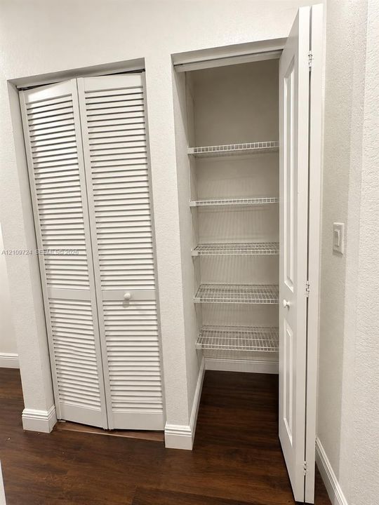 Storage closet featuring white louvered doors, a secondary door with integrated shelving, wire rack shelving units, and wood-finish flooring