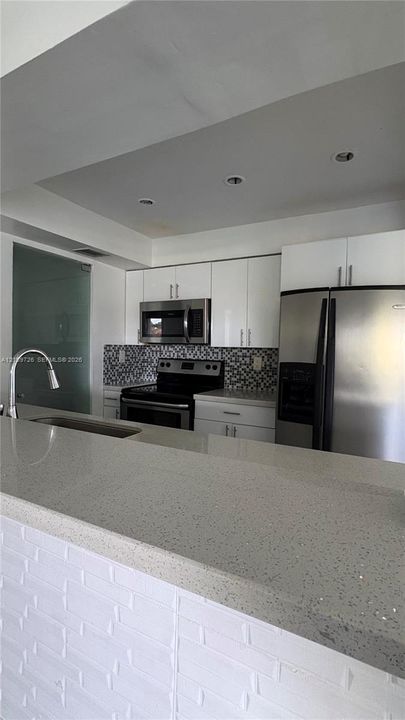 Kitchen featuring white flat-panel cabinetry, quartz countertops, stainless steel appliances, a mosaic tile backsplash, and a white brick-patterned bar surround