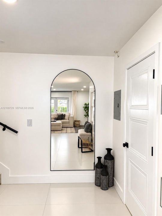 Large arched wall mirror with a matte black frame, light-toned tile flooring, and a multi-panel door with matte black hardware