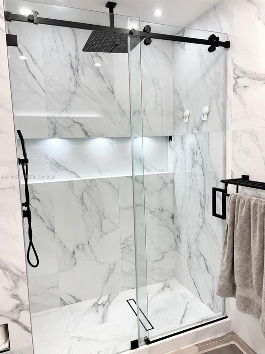 Walk-in shower featuring large-format stone-finish wall and floor tile, glass sliding door enclosure, black rainfall shower head with handheld attachment, and an integrated recessed shelf