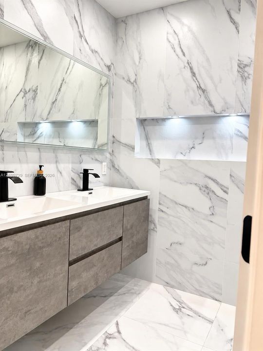 Floating vanity with gray-textured cabinetry, double-sink countertop, matte black fixtures, full-wall stone-patterned tile, and recessed wall niche with integrated lighting
