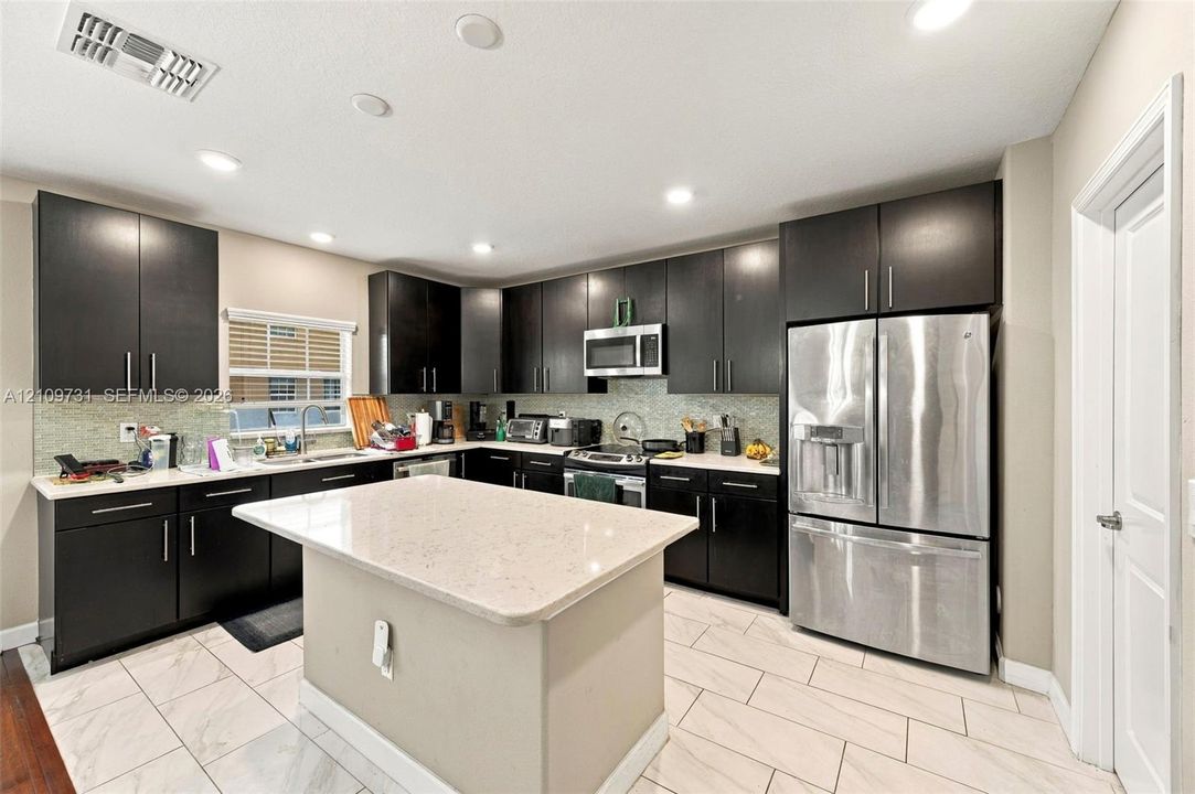 Kitchen featuring dark cabinetry, stone-surface countertops, central island, stainless steel appliances, and large-format tile flooring