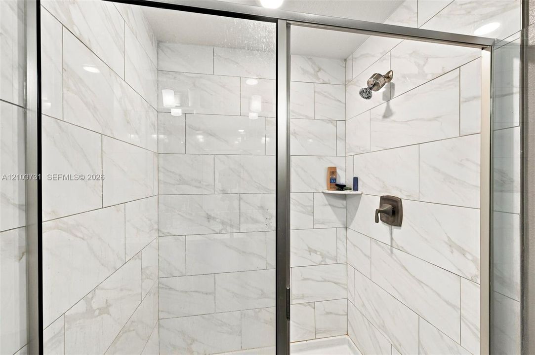 Walk-in shower featuring floor-to-ceiling light-toned tile with subtle veining, framed glass enclosure, built-in corner shelf, and oil-rubbed bronze plumbing fixtures