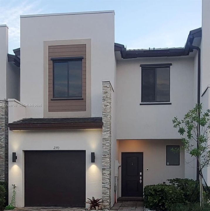 Modern stucco facade featuring horizontal wood-finish siding accents, decorative stone veneer pillars, integrated wall-mounted exterior lighting, and a paved driveway