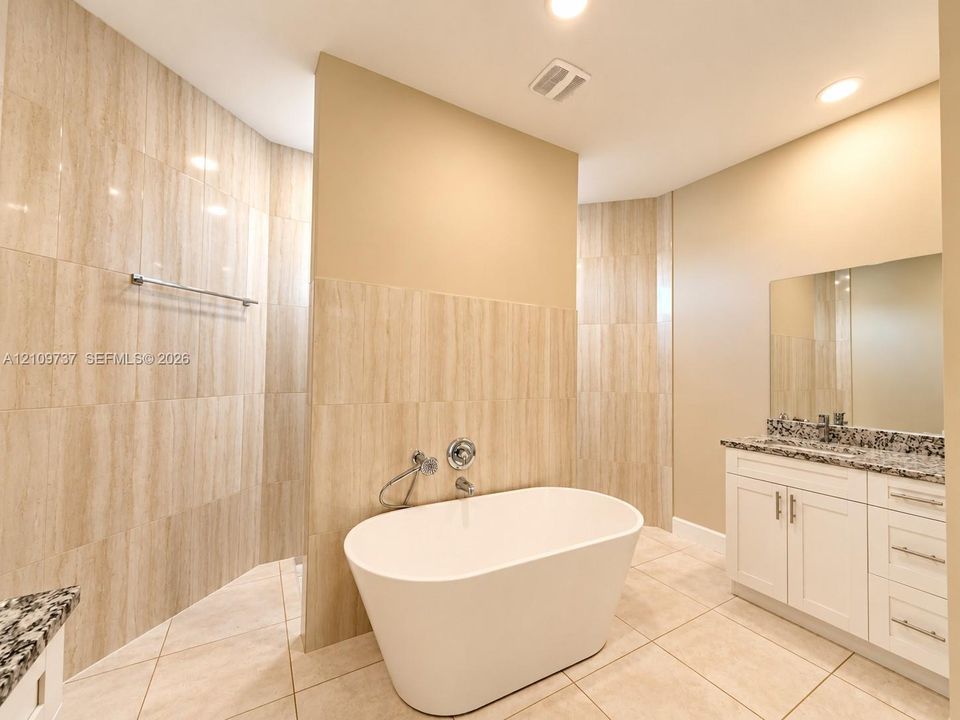 Freestanding soaking tub with a wall-mounted faucet, walk-in shower with floor-to-ceiling tile, vanity with stone-finish countertop, and recessed lighting