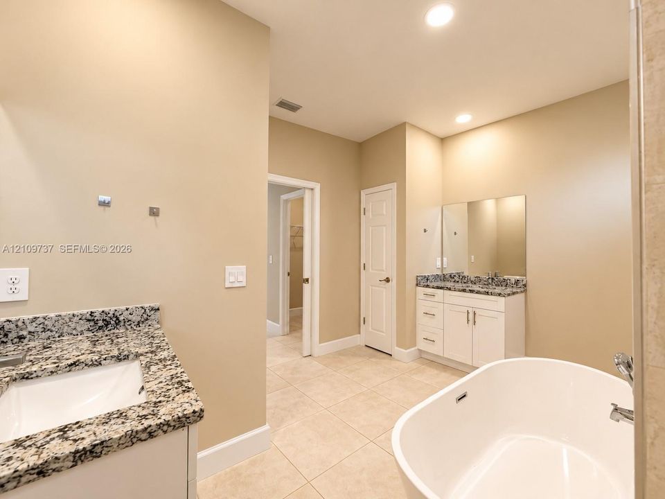Bathroom featuring dual white vanities with stone countertops, a freestanding soaking tub, large-format tile flooring, and recessed lighting