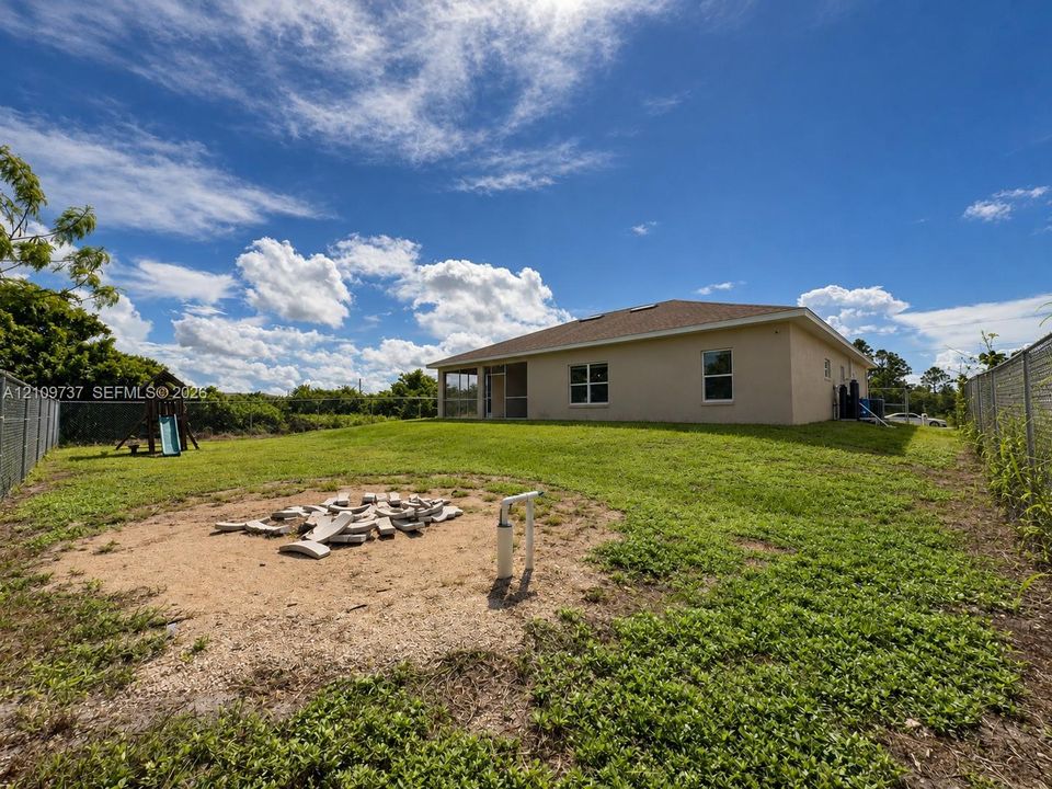 Spacious backyard featuring a level lawn, perimeter chain-link fencing, a dedicated circular gravel fire pit area, and a rear-facing screened-in porch
