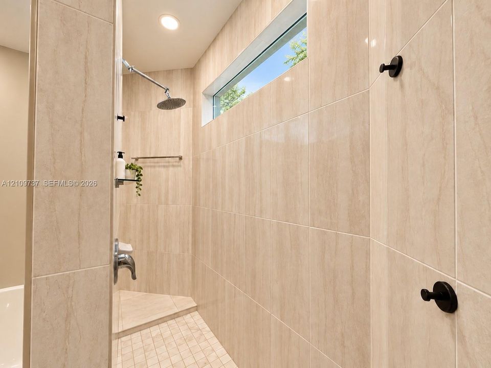 Walk-in shower featuring floor-to-ceiling neutral tile, rainfall showerhead, mosaic tile floor, horizontal transom window, and matte black hardware