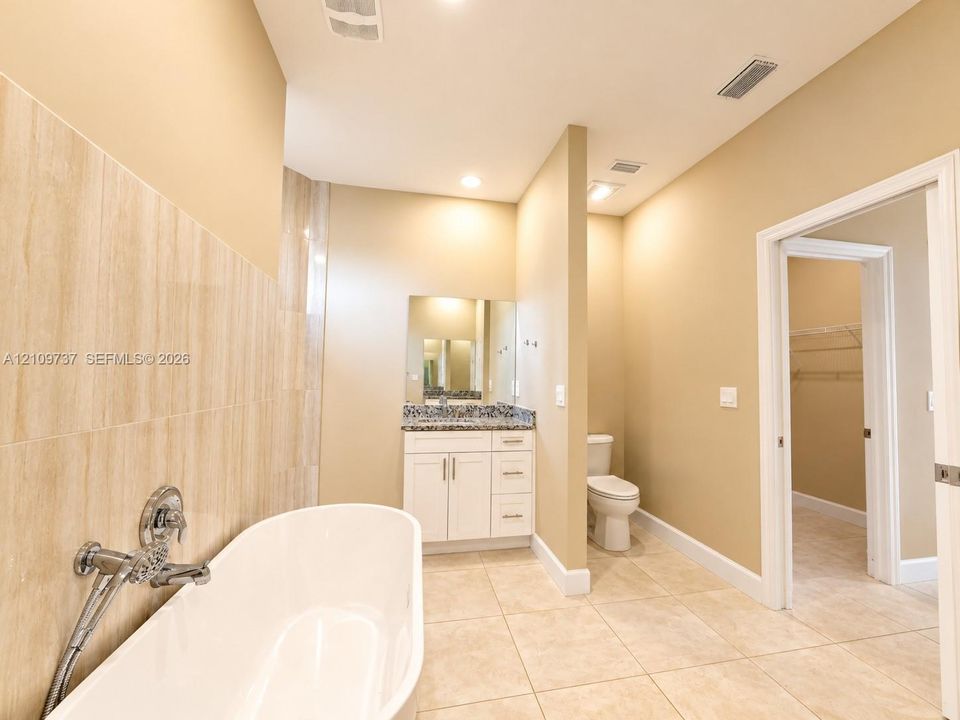 Freestanding soaking tub with wall-mounted chrome hardware, white shaker-style vanity, stone-surfaced countertop, and large-format tile flooring