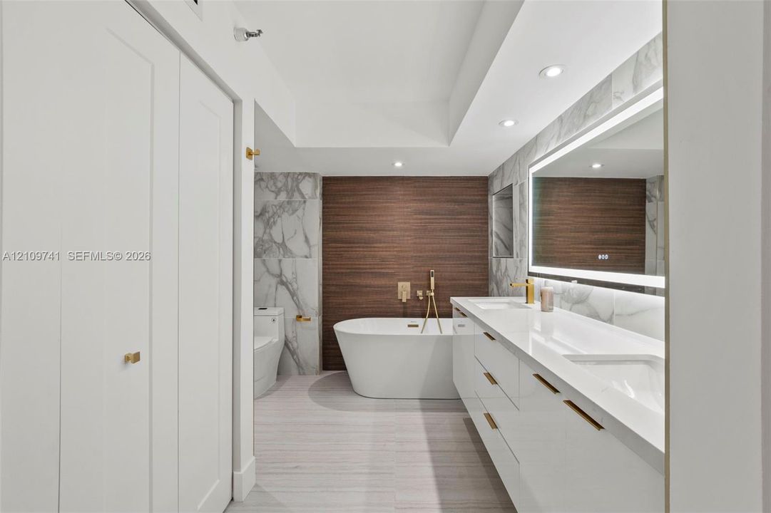Modern bathroom featuring a dual-sink vanity with white cabinetry, freestanding soaking tub with wall-mounted gold-tone hardware, large-format tiled walls, and LED-backlit mirror