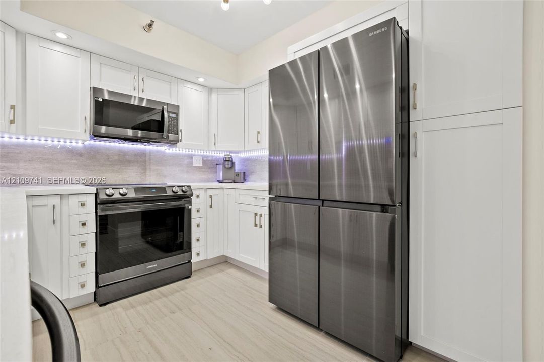 Kitchen featuring white shaker-style cabinetry, stainless steel appliances, stone-surface countertops, under-cabinet lighting, and wood-finish flooring