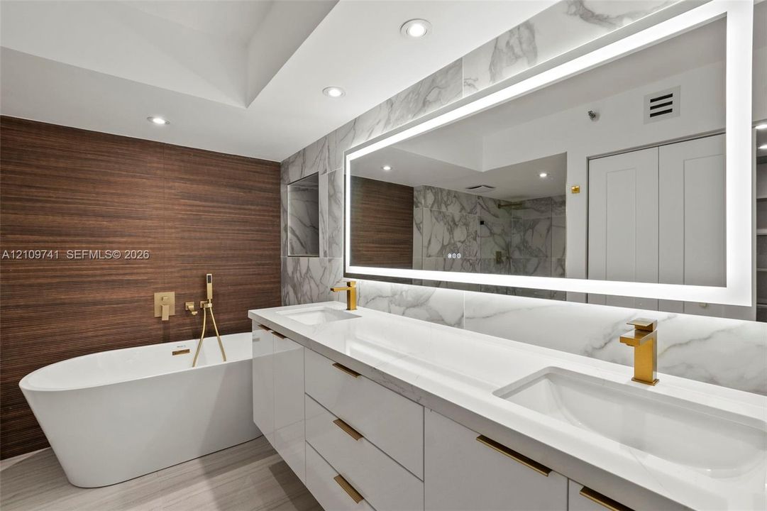 Freestanding soaking tub with wall-mounted gold-toned hardware, white vanity with integrated double sinks, large LED-backlit mirror, wood-finish feature wall, and stone-finish wall tile