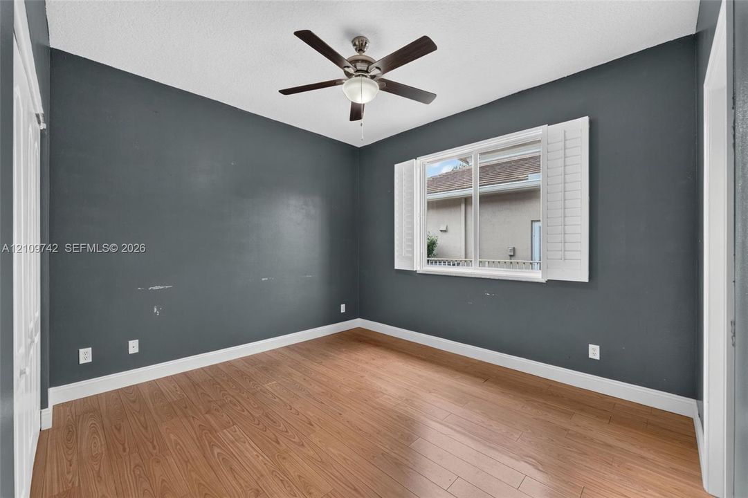 Interior room featuring wood-finish flooring, dark-toned walls, a five-blade ceiling fan with integrated lighting, and plantation-style window shutters