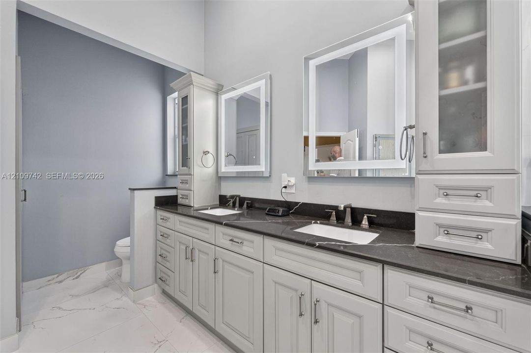 Dual-sink vanity with dark stone countertop, white shaker-style cabinetry, illuminated wall mirrors, and porcelain tile flooring