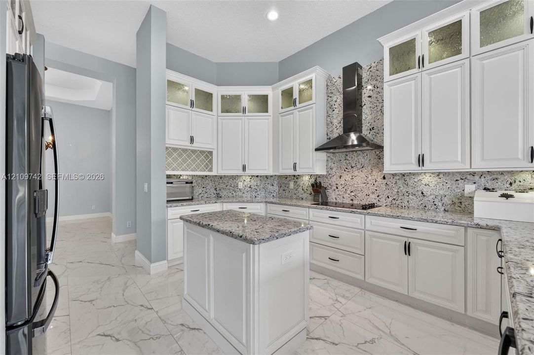 Kitchen featuring white shaker-style cabinetry with glass-panel uppers, patterned stone backsplash, center island, integrated cooktop with stainless steel range hood, and tile flooring