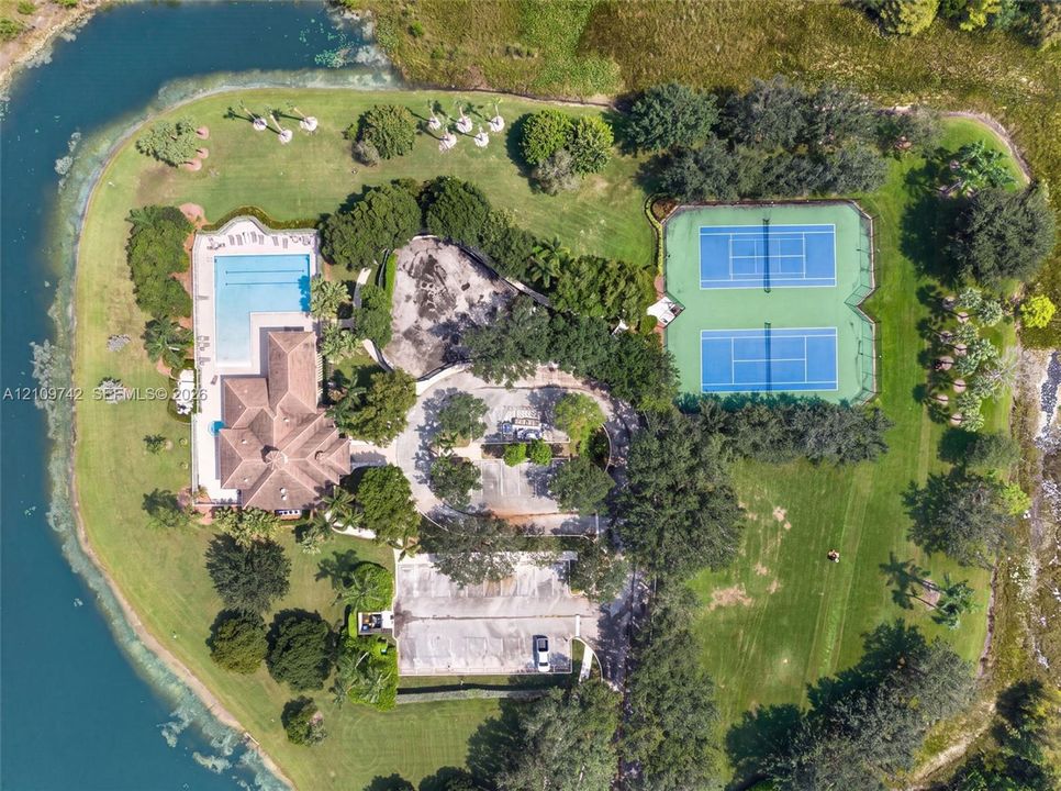Aerial perspective of a multi-acre property featuring a swimming pool, two hard-court tennis courts, expansive manicured lawn, and water frontage