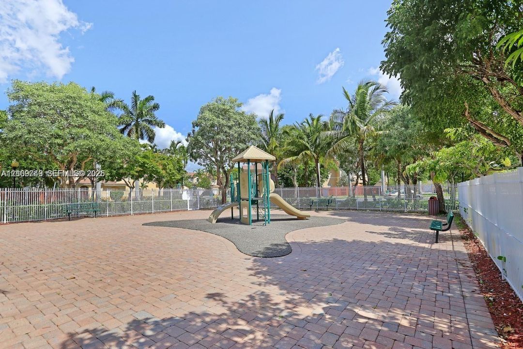 Recreational area featuring paver-stone hardscaping, a multi-component play structure with a molded rubber landing pad, perimeter metal fencing, and mature tropical landscaping