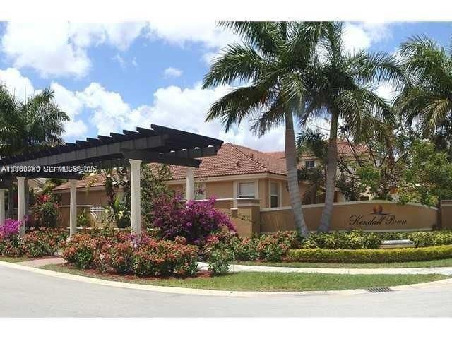 Community entrance featuring a decorative pergola structure, stucco perimeter wall, mature palm trees, and landscaped gardens with flowering shrubs