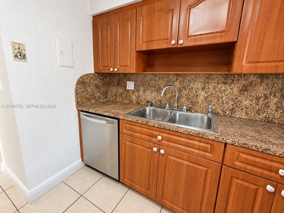 Kitchen area featuring wood-finish cabinetry, solid-surface countertops with an integrated backsplash, stainless steel double-basin sink, and a stainless steel dishwasher