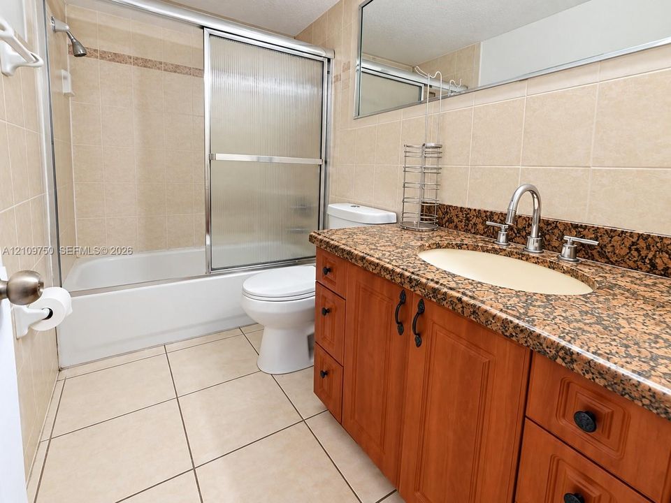 Bathroom featuring a vanity with dark-toned stone-finish countertop, undermount sink, and wood-finish cabinetry, paired with a shower-tub combination, sliding glass enclosure, and ceramic tile flooring