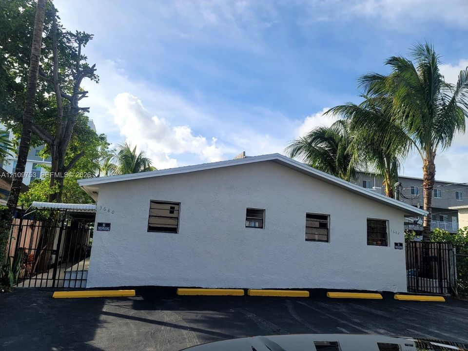 Single-story structure featuring a light-colored textured exterior, multiple jalousie windows, asphalt paving, and metal perimeter fencing