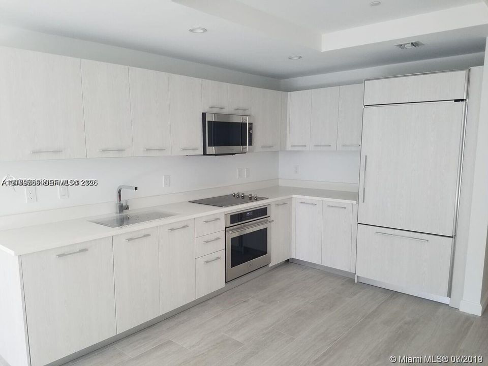 Kitchen featuring light wood-grain cabinetry, solid surface countertops, stainless steel appliances, a flush-mounted cooktop, and wood-finish flooring