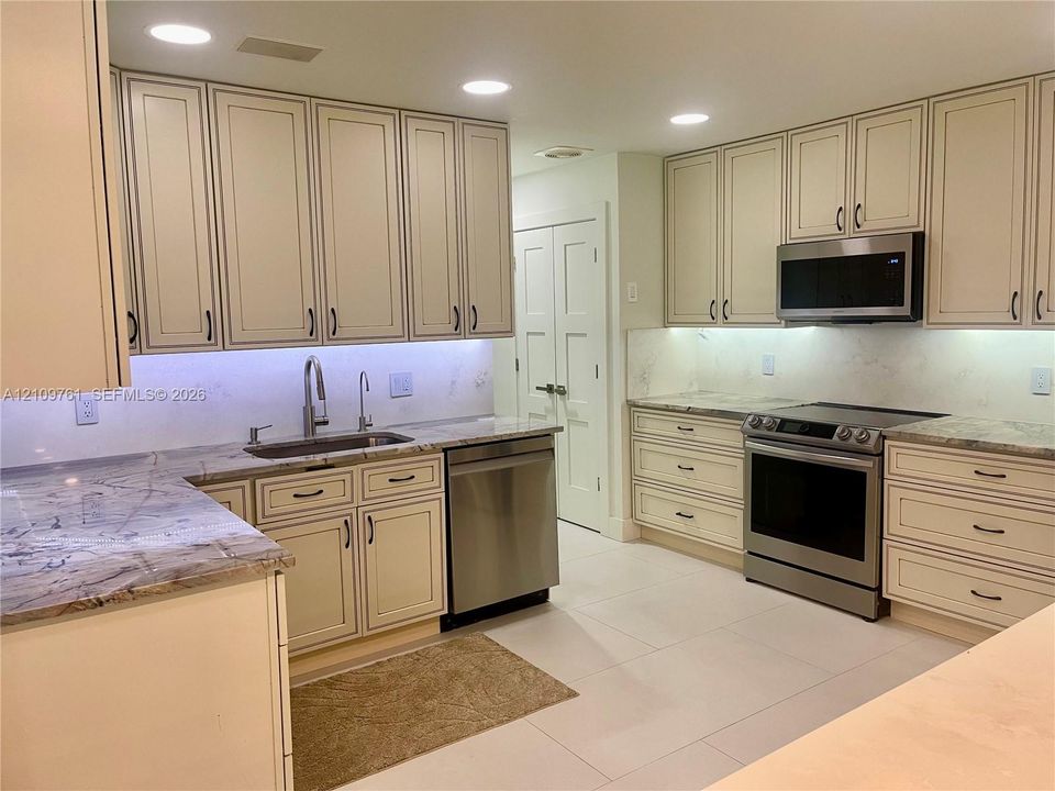 Kitchen featuring off-white cabinetry, stone-finish countertops, stainless steel appliances, under-cabinet lighting, and large-format tile flooring