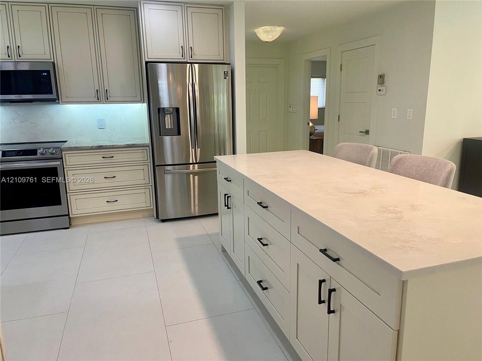 Kitchen featuring an expansive central island with stone-finish countertops, stainless steel appliances, shaker-style cabinetry, and large-format tile flooring