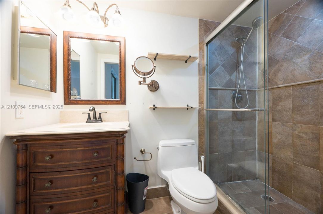 Bathroom featuring a wooden vanity cabinet, stone-surface countertop, shower enclosure with tile walls, and wall-mounted shelving