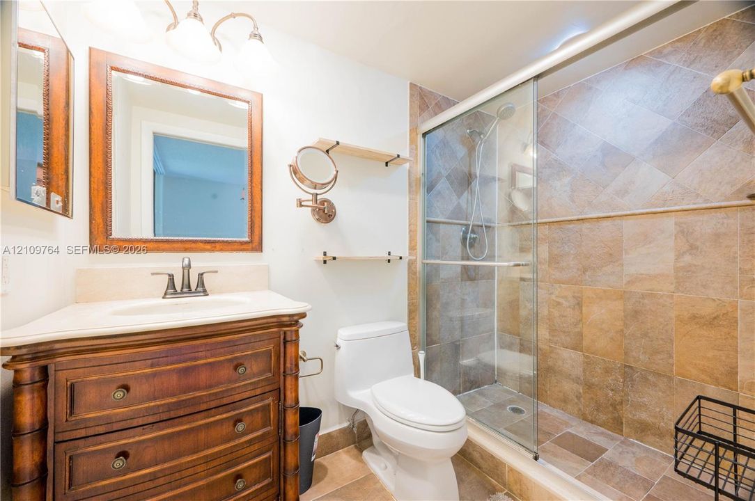 Bathroom featuring a wooden vanity with a stone countertop, framed mirror, wall-mounted shelving, and a tiled shower enclosure with sliding glass doors