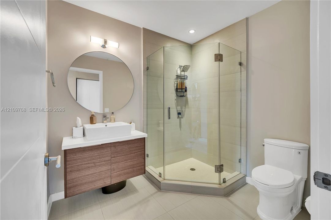 Floating wood-finish vanity with a rectangular vessel sink and white countertop, complemented by a round wall-mounted mirror, brushed-metal light fixture, and a glass-enclosed corner shower with tile surround