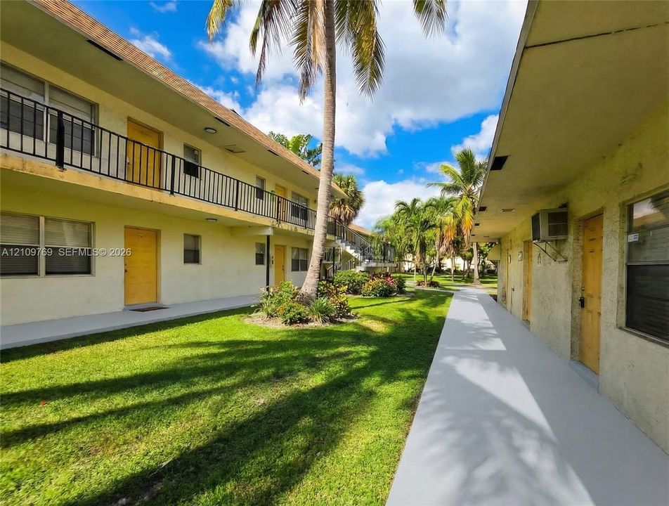 Two-story multi-unit structure featuring stucco exterior, metal balcony railings, concrete walkways, and mature palm trees