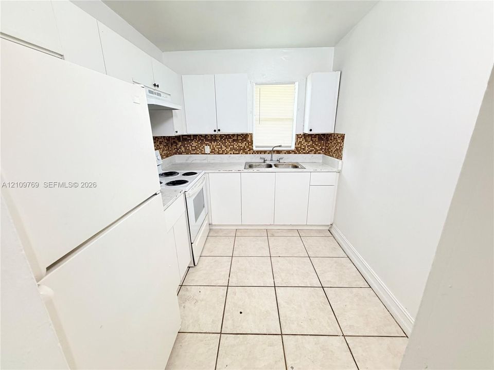 Kitchen featuring white cabinetry, solid-surface countertops, dual-basin sink, mosaic tile backsplash, and light-toned ceramic tile flooring
