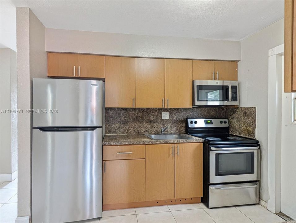 Kitchen area featuring light-finish wood cabinetry, mottled-stone-patterned countertops, stainless steel appliances, a built-in microwave, and light-tone tile flooring