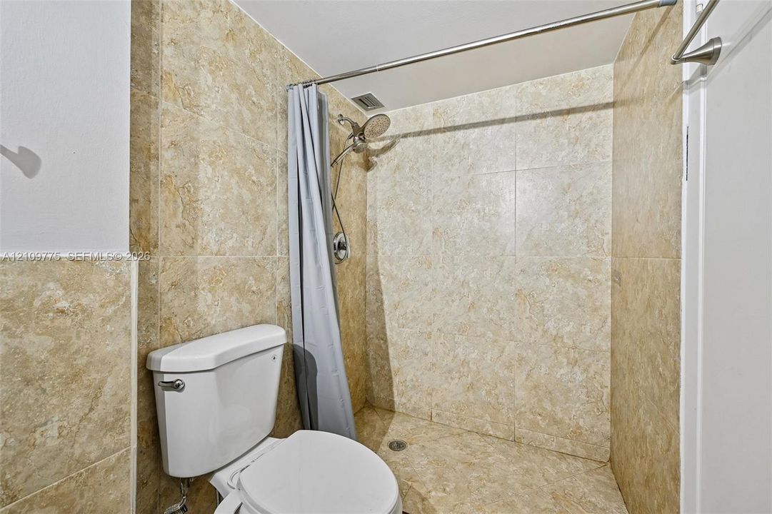 Full-height stone-finish wall tiling, walk-in shower stall with an adjustable-height showerhead, and a high-profile white porcelain toilet