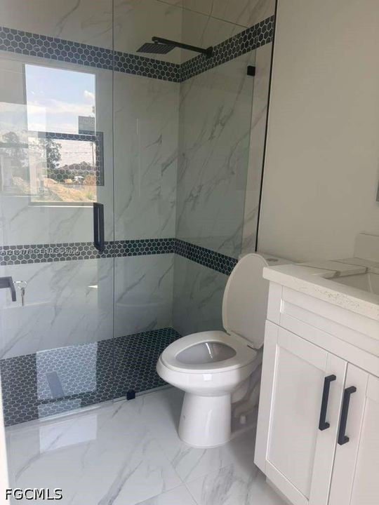 Walk-in shower featuring large-format tile surrounds with black hexagonal accent bands, a frameless glass enclosure, a matte black rain shower head, and a matching stone-topped vanity with black hardware