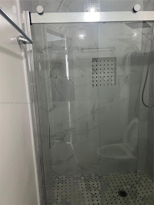 Walk-in shower featuring large-format stone-look wall tile, glass sliding door, built-in recessed tiled niche, rainfall shower head, and hexagonal mosaic floor tile