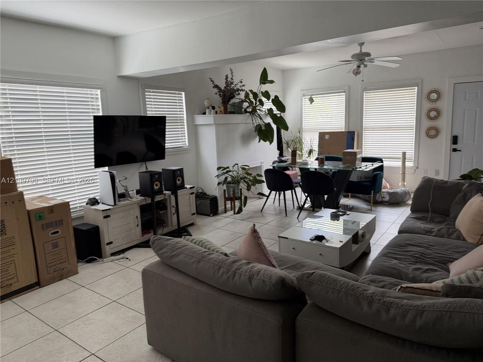 Living area featuring large format neutral tile flooring, neutral wall tones, multiple windows with integrated blinds, a central ceiling fan, and a prominent decorative fireplace mantle