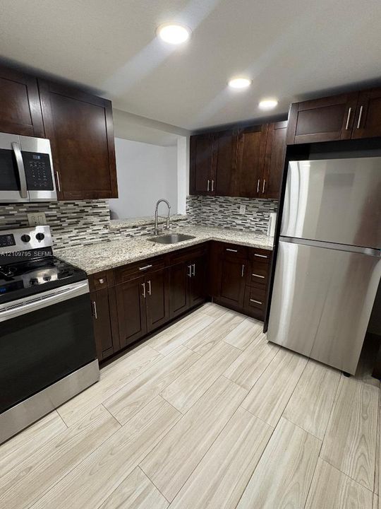 Kitchen featuring dark wood cabinetry, stainless steel appliances, multi-tonal tile backsplash, stone-finish countertops, and wood-finish flooring