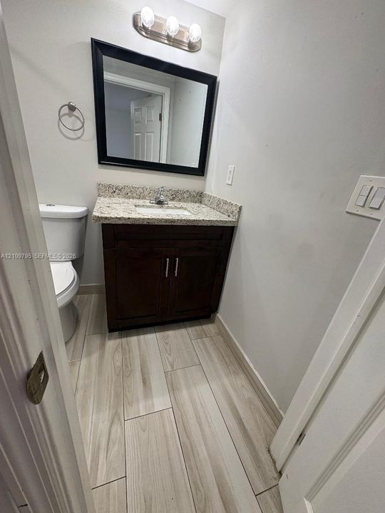 Bathroom featuring wood-finish tile flooring, a dark wood vanity with a stone countertop, a framed rectangular mirror, and a three-bulb light fixture