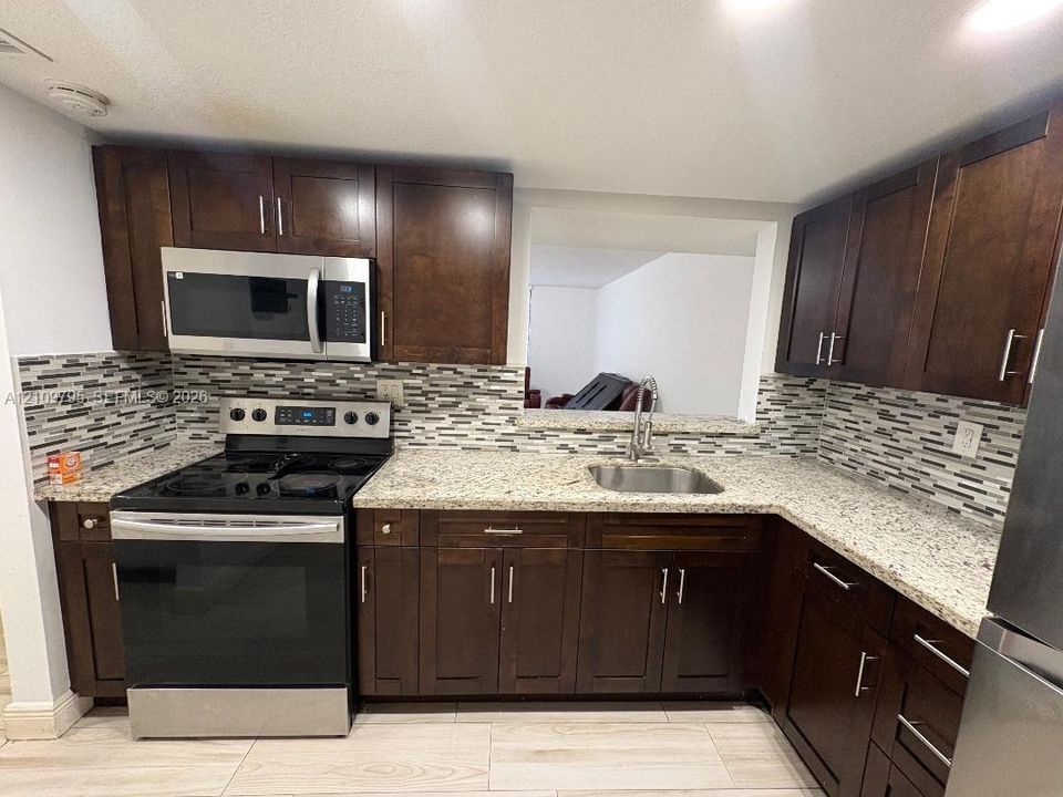 Kitchen featuring dark wood cabinetry, stone-finish countertops, stainless steel appliances, multi-toned tile backsplash, and wood-finish tile flooring