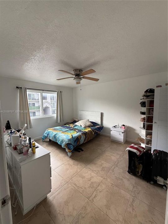 Master bedroom interior featuring large-format tile flooring, a multi-blade ceiling fan with light fixture, and a single-pane sliding window with white louvered closet doors