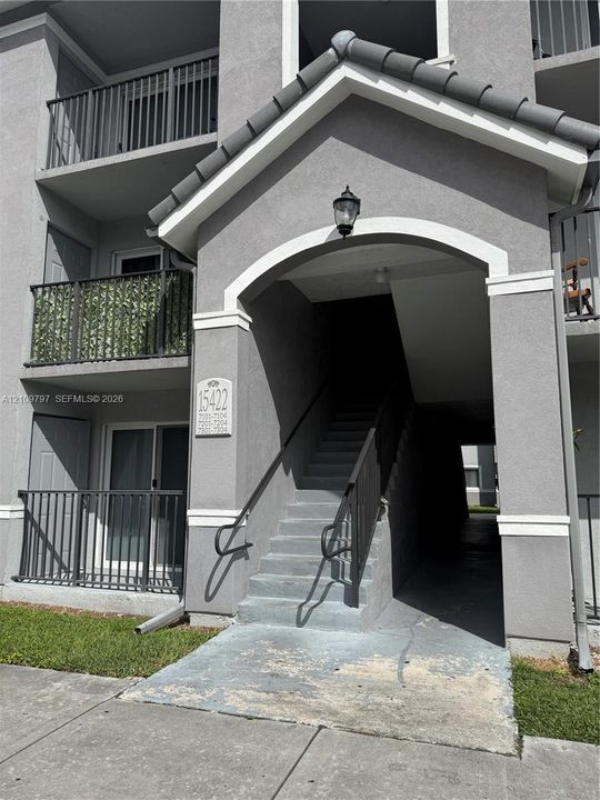 Stucco exterior featuring a gabled entryway, metal balcony railings, concrete stairs, and exterior wall-mounted lighting