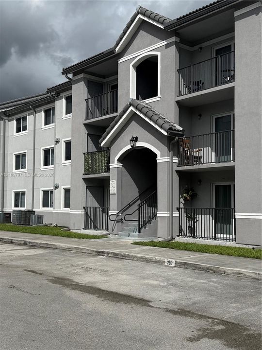 Multi-story exterior featuring a grey stucco facade, dark shingled roof, arched entryway, and private balconies with black metal railings