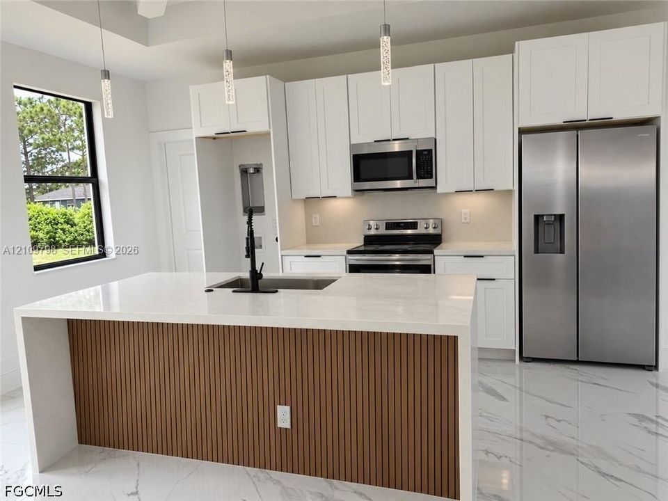 Kitchen featuring white shaker-style cabinetry, quartz countertops, a central island with a slatted wood-finish base, stainless steel appliances, and large-format tile flooring