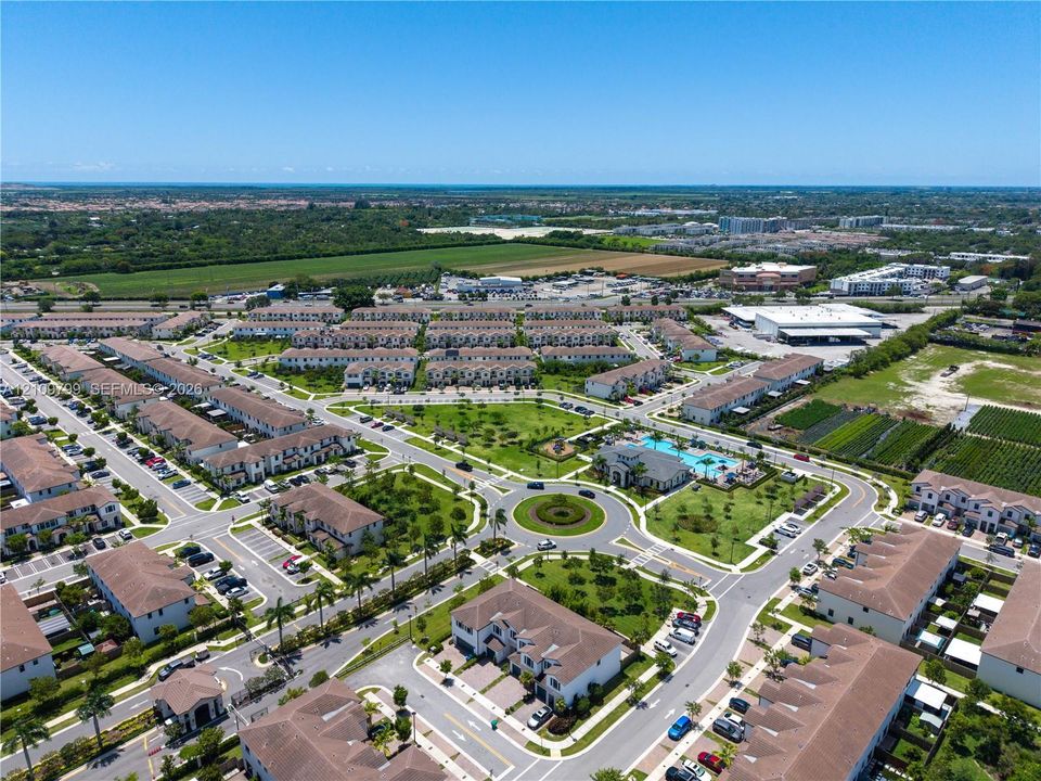 Aerial perspective of a multi-unit residential community featuring paved roads, a central traffic circle, manicured green spaces, and a swimming pool facility