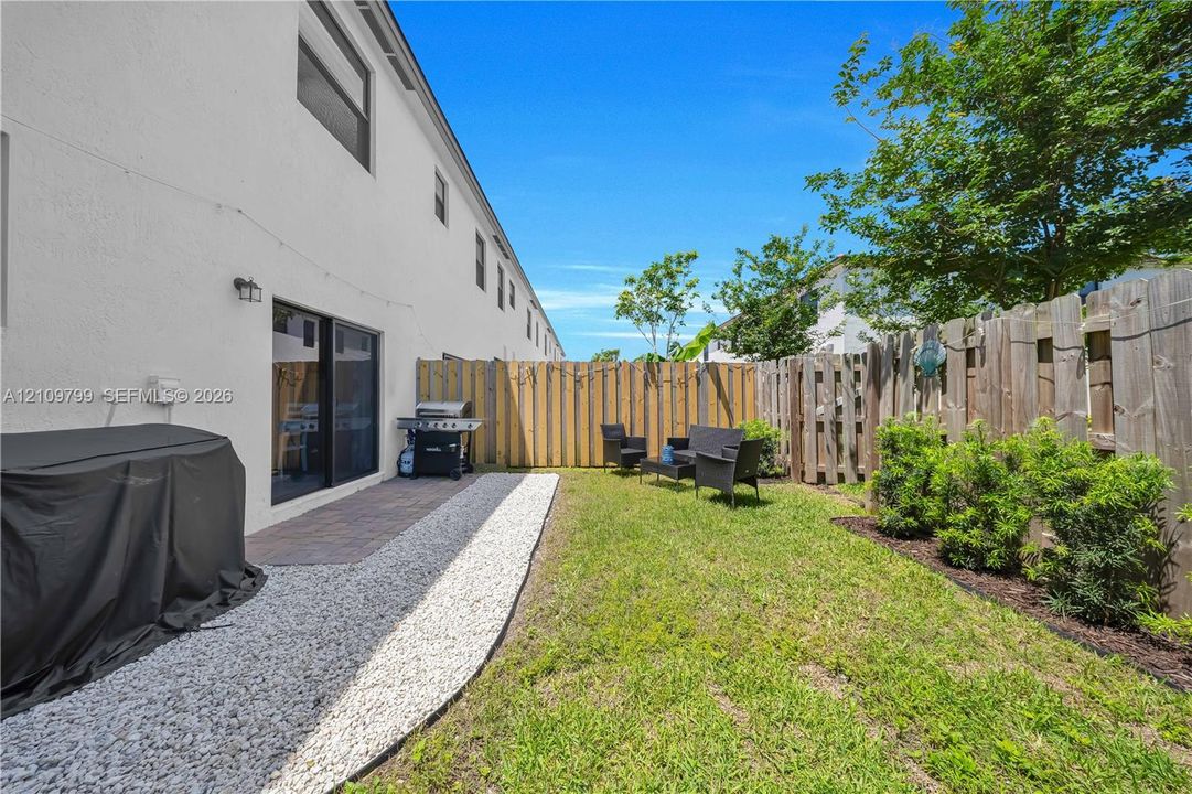 Fenced backyard featuring a paver patio, stone-accented ground cover, established landscaping, and exterior wall-mounted lighting