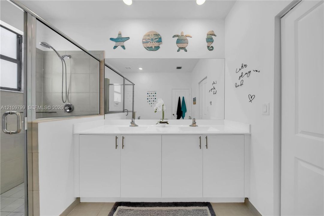 Dual-sink vanity with white cabinetry, a solid-surface countertop, and a full-width mirror, paired with a glass-enclosed shower featuring tile surrounds and a wall-mounted showerhead